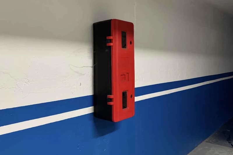 Rotes schwarzes Wandgehäuse an weiß-blauer Wand mit zwei blauen Streifen im Parkhaus.