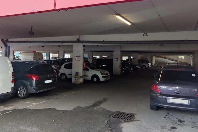 Überdachtes Parkhaus mit abgestellten Autos, Betonstützen, Deckenbeleuchtung und Auffahrt im Hintergrund.