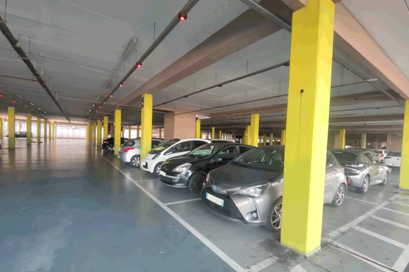 Überdachtes Parkhaus mit gelben Stützen und nebeneinander geparkten Autos, rote Deckenlampen sichtbar.