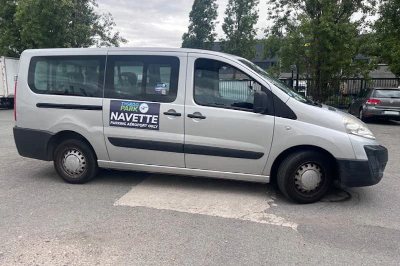 Silberner Shuttle-Van mit Tygroo Park Navette Aufkleber, geparkt auf Asphalt vor Zaun und Bäumen.