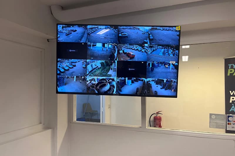 Wandmonitor zeigt mehrere CCTV-Aufnahmen eines Parkplatzes, Glasfenster und ein Feuerlöscher im Raum.