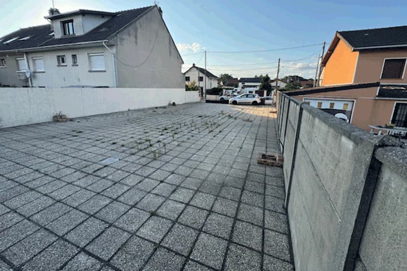Gepflasterte Parkfläche mit quadratischen Platten und einzelnen Unkrautpflanzen, umgeben von niedrigen Mauern und Wohnhäusern.
