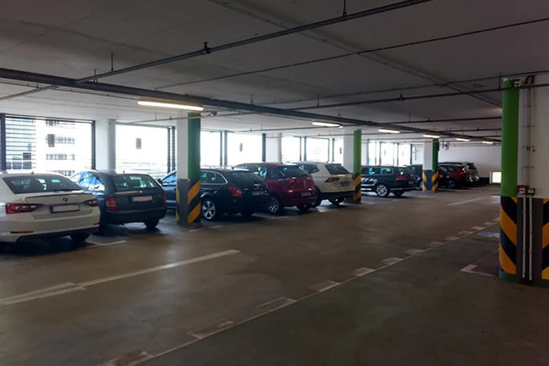 Überdachte Parkebene mit geparkten Autos, grün lackierten Säulen mit gelb-schwarzen Warnstreifen und seitlichen Fenstern.