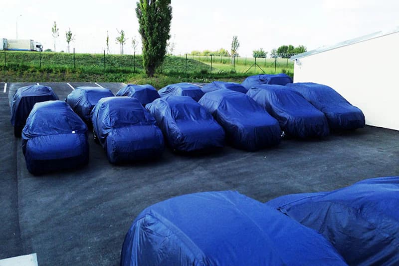 Außenparkplatz mit mehreren Autos unter blauen Abdeckhauben, umzäunter Hintergrund und Grünfläche am Rand.