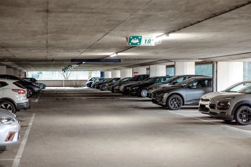 Überdachte Parkhalle mit Reihen geparkter Fahrzeuge, Betonstützen, Deckenleuchten und grünem Parkfeldschild 18 sichtbar.