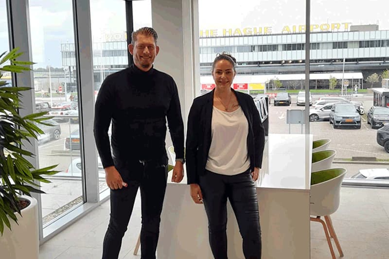 Zwei Personen stehen in einem hellen Büro vor großen Fenstern mit Blick auf das Flughafenterminal und geparkte Autos.