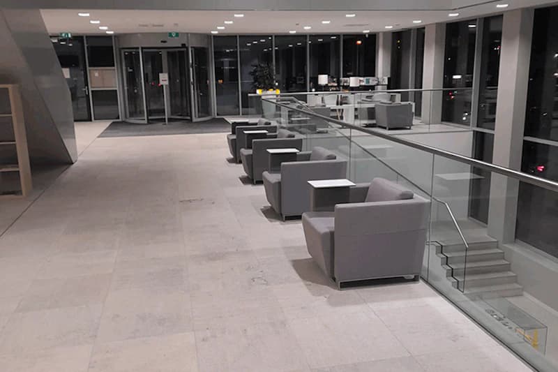 Leere Sitzreihe mit grauen Sesseln und kleinen Tischen an Glasbrüstung in modernem Flughafenfoyer.