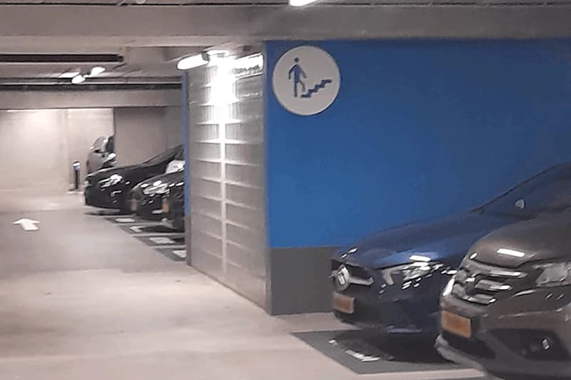 Parkende Autos in einer beleuchteten Tiefgarage, blauer Wandbereich mit Schild für Fußgänger und Pfeil auf dem Boden.