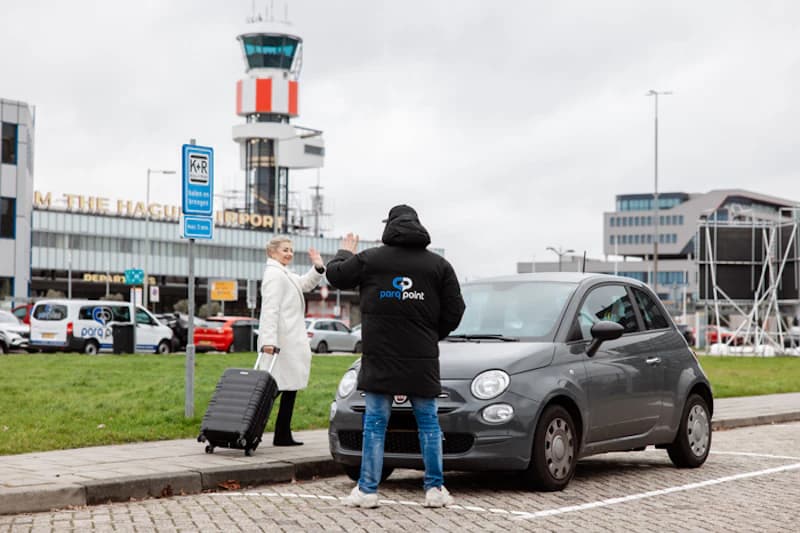 Mitarbeiter mit Parq Point Jacke übergibt Schlüssel an Reisende mit Koffer vor grauem Kleinwagen am Flughafen.
