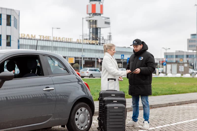 Frau übergibt Schlüssel an Parkservice-Mitarbeiter neben Auto und zwei Koffern vor Rotterdam The Hague Airport.