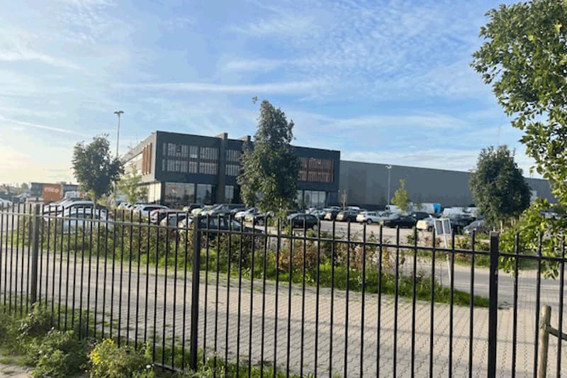 Metallzaun im Vordergrund, dahinter Parkplatz mit zahlreichen Autos und einem zweigeschossigen Gebäude unter blauem Himmel.