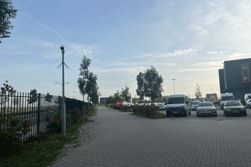 Gezäunter, beleuchteter Parkbereich mit gepflasterter Einfahrt, geparkten Autos und Überwachungskamera an einem Mast.