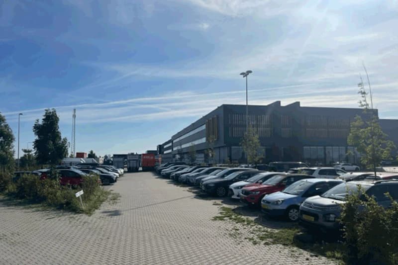 Großer gepflasterter Parkplatz mit Reihen geparkter Autos, Laterne und mehrstöckigem Gebäudekomplex im Hintergrund.