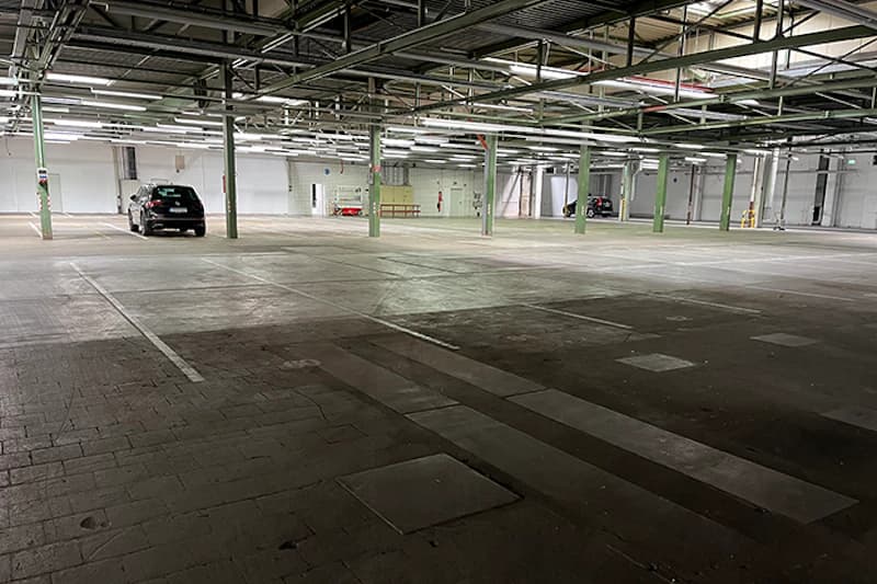 Große leere Parkhalle mit beleuchteten Deckenbalken, grünen Stützen und wenigen geparkten Autos.