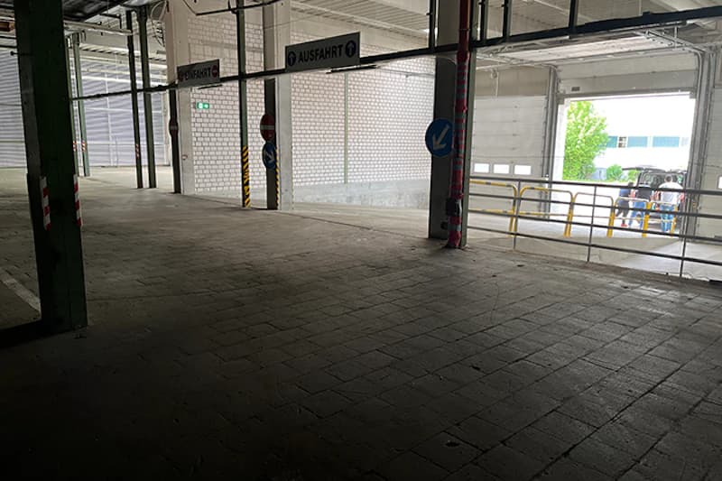 Innenraum einer Parkhalle mit Pfeilschildern Ausfahrt, Betonboden, Stützen und offener Rampe.