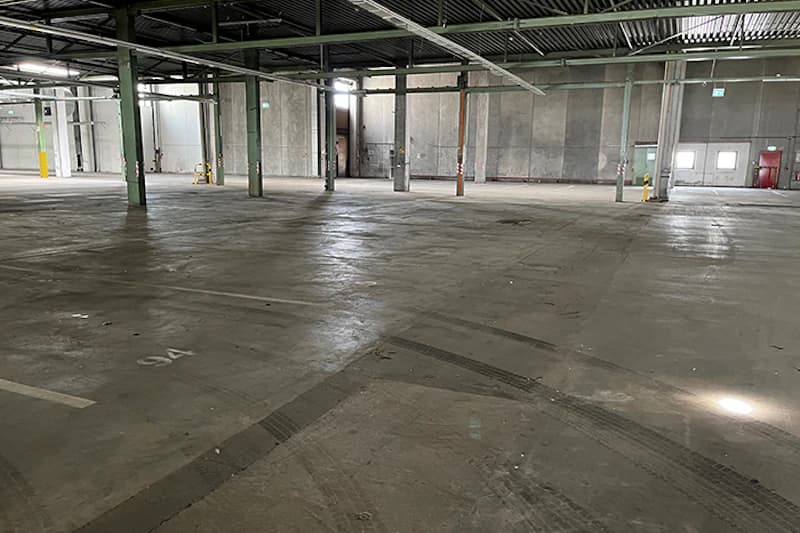Leere Parkhalle mit Betonboden, Stützsäulen, Deckeninstallationen und markierten Parkflächen.
