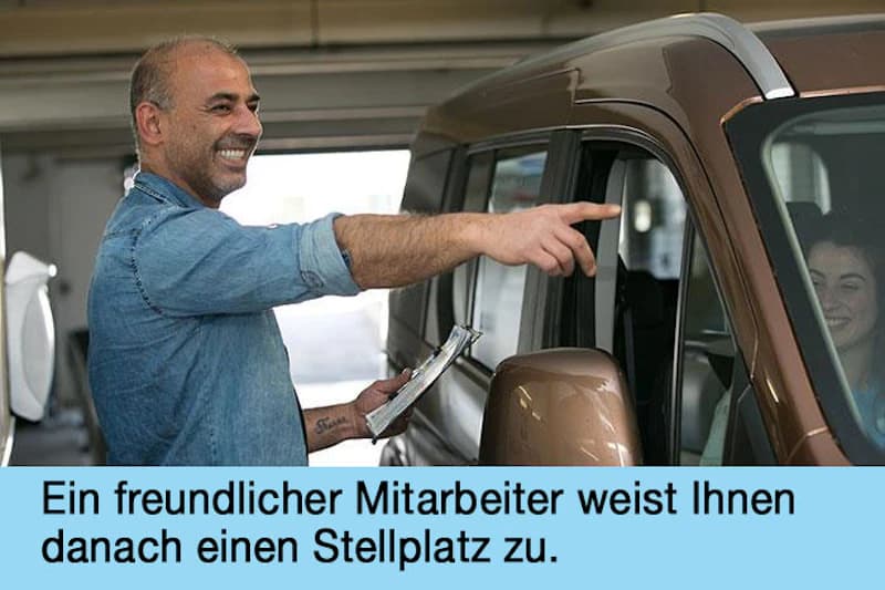 Mann mit Klemmbrett zeigt lächelnd auf ein Fahrzeug in einer Parkgarage, Fahrerin im Auto lächelnd.