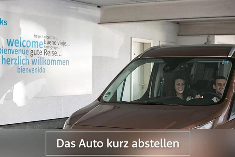 Braunes Auto mit zwei Personen auf überdachtem Parkdeck vor Wand mit mehrsprachigem Willkommensschriftzug.