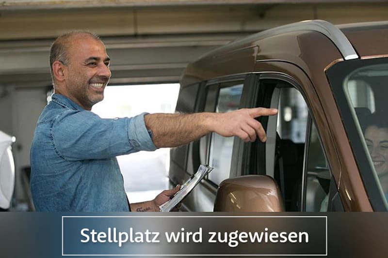 Mann mit Klemmbrett zeigt lächelnd auf ein Auto in einer Parkgarage; Fahrerin im Fahrzeug und Schriftzug Stellplatz wird zugewiesen.