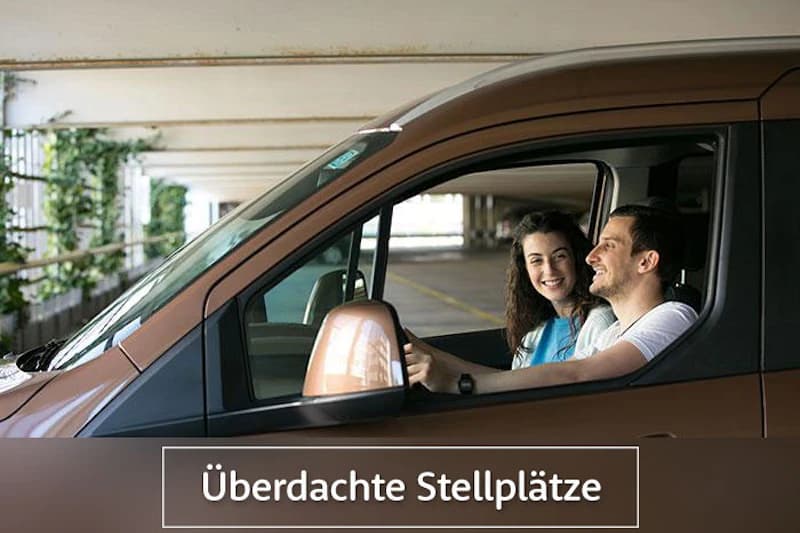 Paar sitzt lächelnd in einem Auto in einem überdachten Parkhaus, seitlicher Spiegel und Parkreihen sichtbar.