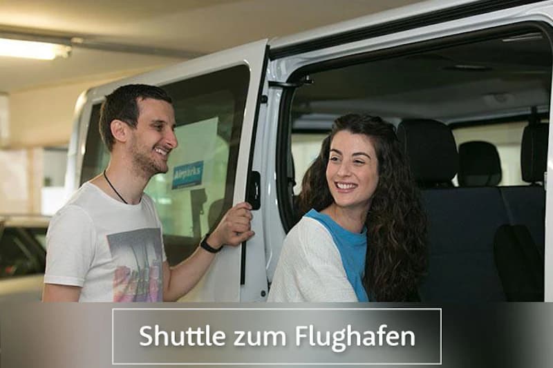 Mann öffnet Tür eines weißen Kleinbusses in Parkhaus, Frau sitzt lächelnd auf dem Rücksitz.
