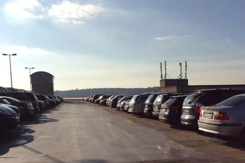 Oberes Parkdeck mit zwei Reihen geparkter Autos, freier Fahrspur, Laternen und kleinem Technikgebäude am Horizont.