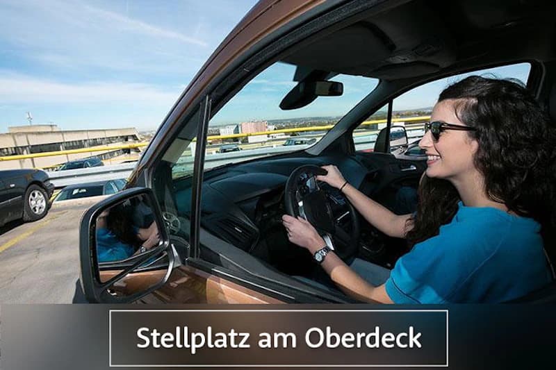 Frau sitzt lächelnd am Steuer in geparktem Auto auf dem obersten Parkdeck mit weiteren Fahrzeugen im Hintergrund.