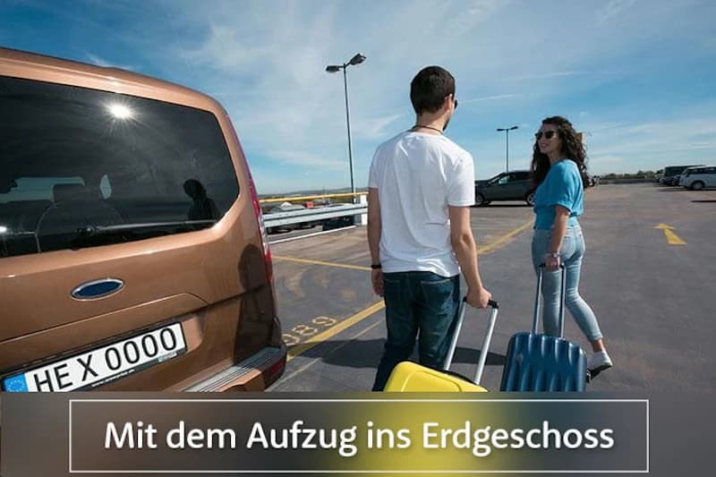 Zwei Personen mit Rollkoffern auf Parkdeck neben geparktem Wagen; Schriftzug Mit dem Aufzug ins Erdgeschoss sichtbar.