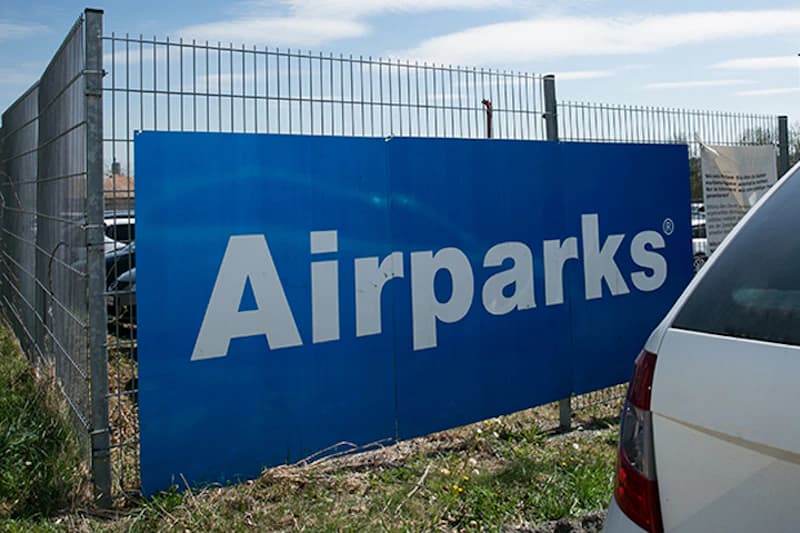 Großes blaues Schild mit weißer Aufschrift Airparks an Drahtzaun neben geparktem weißen Auto.