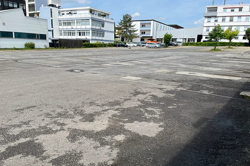 Weitläufiger asphaltierter Parkplatz mit verblassten Stellplatzmarkierungen, wenigen Autos und Gebäuden im Hintergrund.