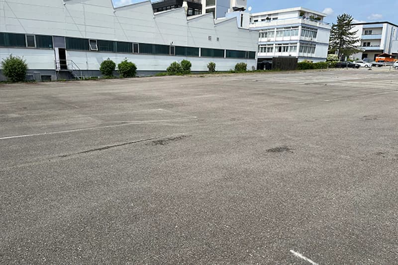 Großer leerer Asphaltparkplatz mit schwachen Parklinien, Industriegebäude und vereinzelten Autos im Hintergrund.