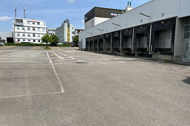 Leerer gepflasterter Hof vor Lagerhalle mit Rampen nummeriert 5 bis 9 und Bürogebäuden im Hintergrund.