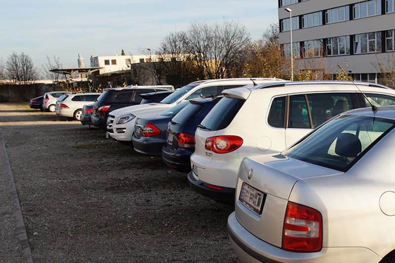 Mehrere nebeneinander geparkte Autos auf Schotterparkplatz neben Bürogebäude und kahlen Bäumen.