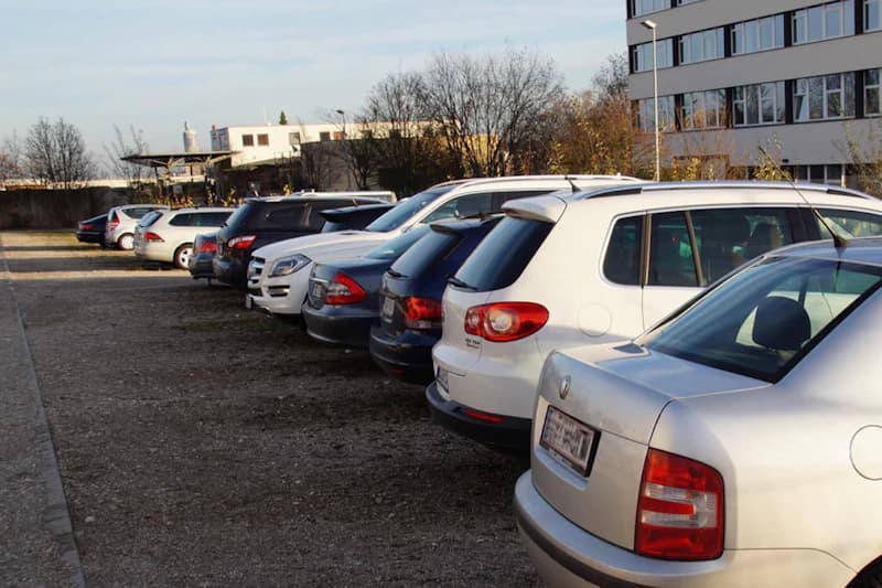 Reihe geparkter Autos auf einem kiesigen Parkplatz neben einem Bürogebäude bei Tageslicht.