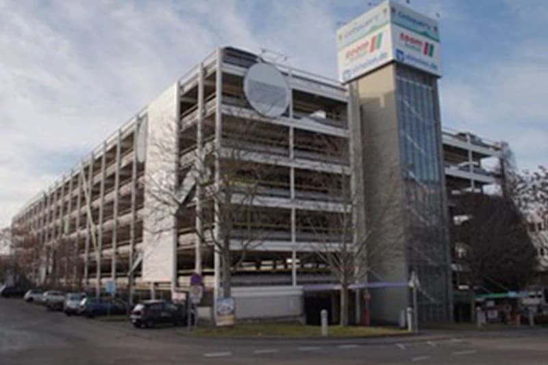 Mehrgeschossiges Parkhaus mit offenem Stahlbetonraster, Aufzugsturm und geparkten Autos an der Straße.