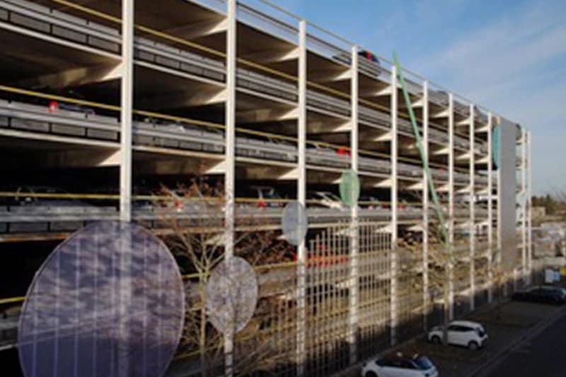 Mehrstöckiges Parkhaus mit offenen Betonetagen und geparkten Autos, dekorative Stelen mit runden Platten im Vordergrund.