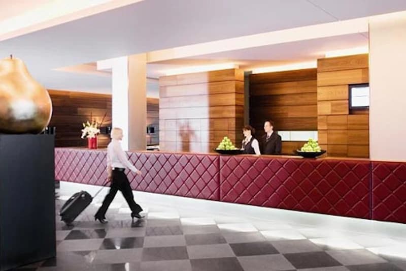 Hotelrezeption mit roter gesteppten Theke, zwei Empfangsmitarbeitenden, vorbeigehendem Gast mit Rollkoffer und großer Dekoskulptur.
