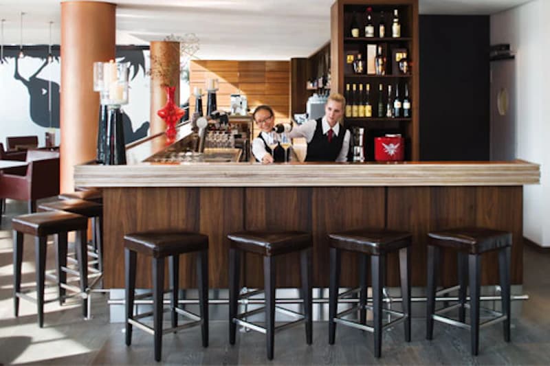 Bar mit Holztresen, fünf Hocker davor und zwei Barkeeperinnen, Regale mit Flaschen im Hintergrund.