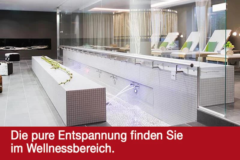 Wellnessbereich mit langem gefliestem Wasserbecken, sprudelnden Massagedüsen, Liegestühlen und Glaswand.
