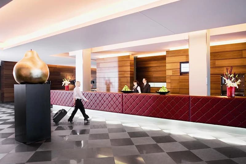Hotellobby mit langer roten Rezeption, zwei Mitarbeitenden hinter dem Tresen und Gast mit Rollkoffer, große goldene Birne.
