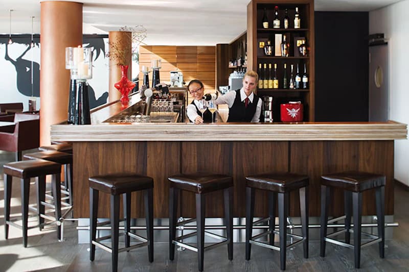 Hotelbar mit Tresen, fünf Hockern und zwei Mitarbeiterinnen, die Getränke einschenken.