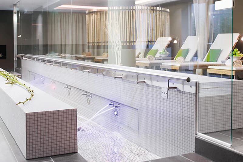 Innenbereich eines Wellnessbereichs mit gefliestem Wasserbecken, seitlichen Wasserdüsen und Liegen hinter Glas.