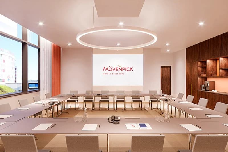 Konferenzraum mit U-förmigen Tischen, Stühlen, Leinwand mit Mövenpick-Logo und runder Deckenleuchte.