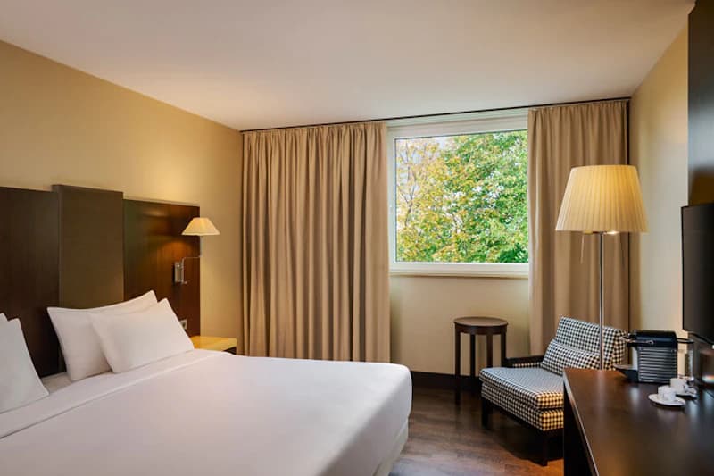 Doppelzimmer mit großem Bett, Kopfteil, Nachttischlampe, Sessel, Schreibtisch mit Kaffeemaschine und Fensterblick auf Bäume.