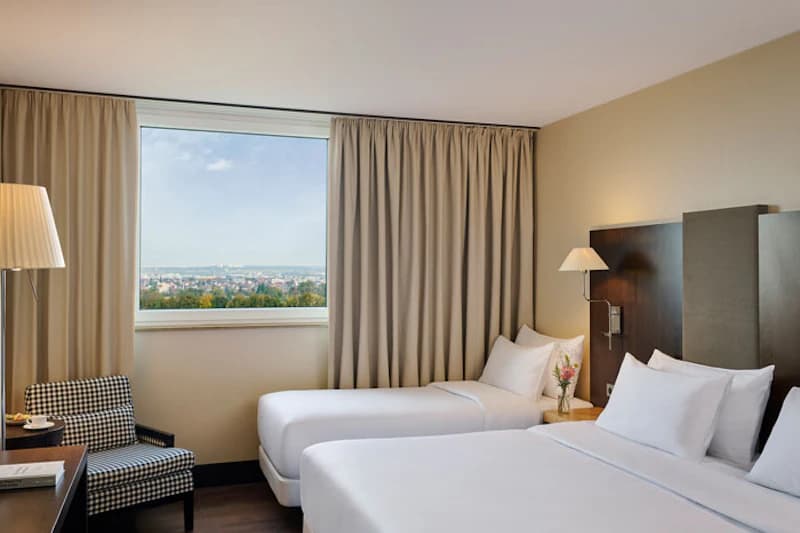 Helles Hotelzimmer mit zwei Einzelbetten, Nachttisch, Leselampen, Sessel und Fenster mit Stadtblick.