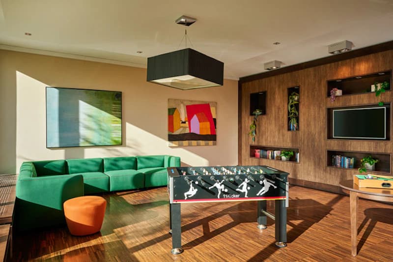 Hotellounge mit grünem Ecksofa, orangefarbener Hocker und Tischkicker vor Wandregal mit Fernseher und Pflanzen.
