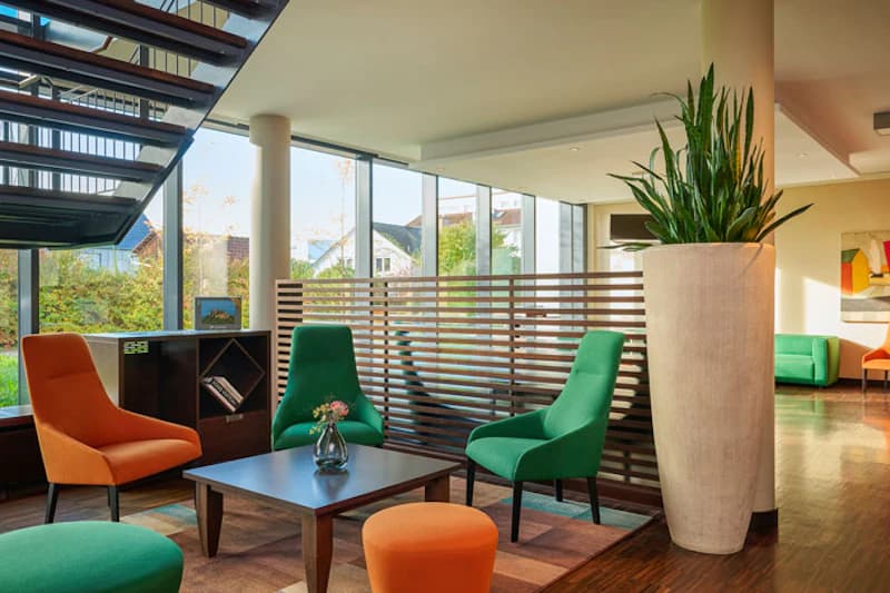 Lobby mit bunten Sesseln, Holztisch, Raumteiler und großem Pflanzenkübel.