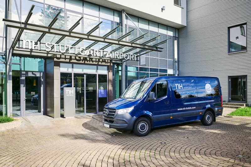 Hoteleingang mit Glasfassade und Vordach mit Schriftzug NH Stuttgart Airport sowie blaues Shuttlefahrzeug davor.