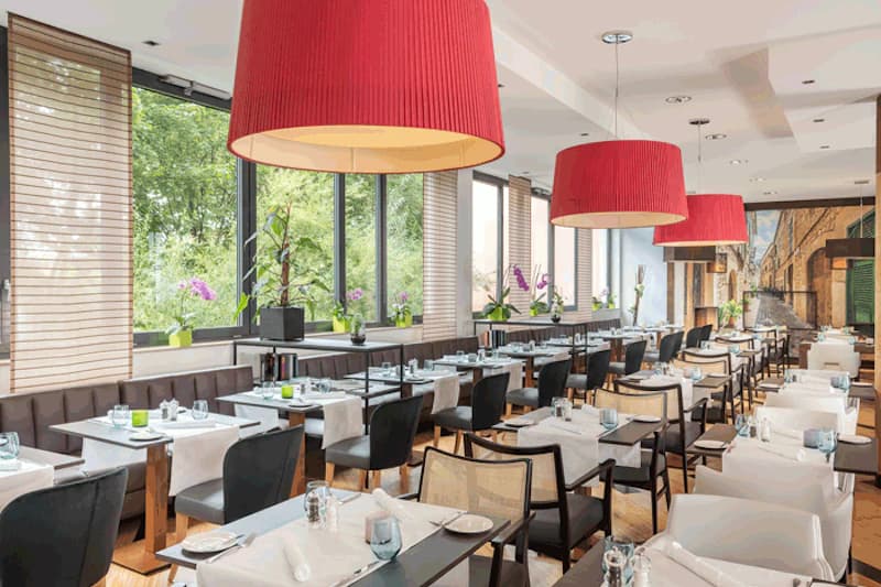 Hoteleigenes Restaurant mit gedeckten Tischen, roten Hängeleuchten und Fenstern zur grünen Außenseite.