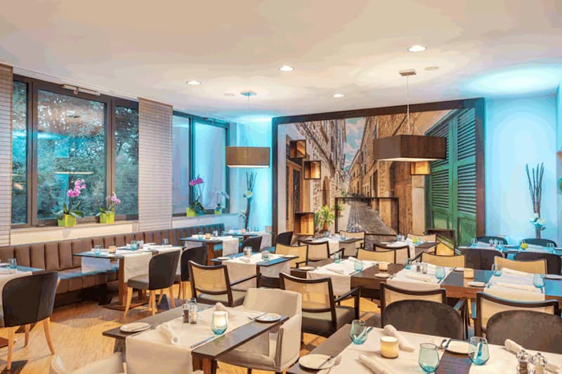 Hotelrestaurant mit gedeckten Tischen, Sitzbänken, blauen Lichtakzenten und großem Wandbild einer Gasse.
